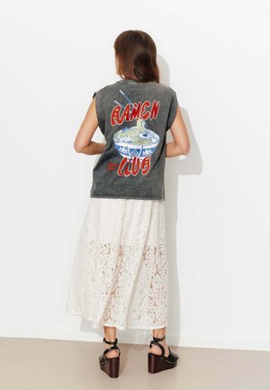 Femme aux longs cheveux portant un haut noir sans manches avec l'inscription « Ramen Club » et un motif de bol de nouilles, une jupe en dentelle blanche et des sandales à plateforme noires.