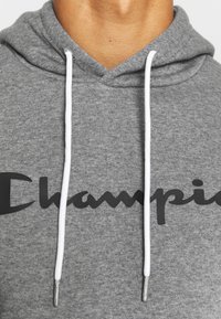 Felpa grigia in cotone con logo "Champion" nero, caratterizzata da un collo a V e cordini bianchi con puntali metallici. Tessuto texturizzato.