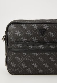 Black Guess crossbody bag with gray monogram pattern and front zip pocket, featuring a triangular Guess logo plaque.