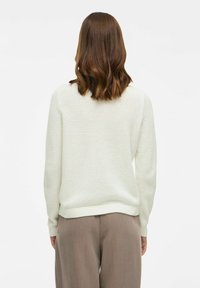 Suéter blanco texturizado con cuello redondo, mangas largas y dobladillo acanalado, combinado con pantalones beige de pierna ancha.