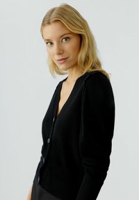Schwarze Strickjacke mit V-Ausschnitt, Knopfverschluss und gerippten Bündchen. Weicher Stoff mit gerafften Details an den Schultern.