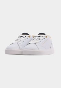 Ζευγάρι λευκά αθλητικά παπούτσια χαμηλής κοπής με μαύρη γλώσσα, λευκά κορδόνια, χρυσά ρέλια και λογότυπο Tommy Hilfiger στο πλάι, πάνω σε λευκό φόντο.