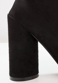 Chaussure à talon haut en daim noir avec un talon épais et carré, posée sur une surface blanche.