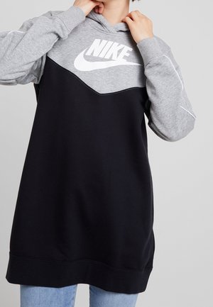 Osoba ubrana w długi czarno-szary sweter Nike z białym logo Nike, poprawiająca kołnierz, w połączeniu z niebieskimi jeansami.