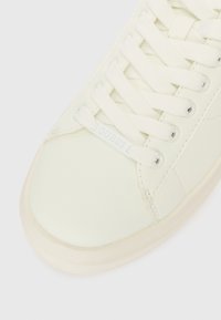 Detailní záběr na bílou tenisku s bílými tkaničkami a značkovým štítkem "GUESS" na bílém pozadí.