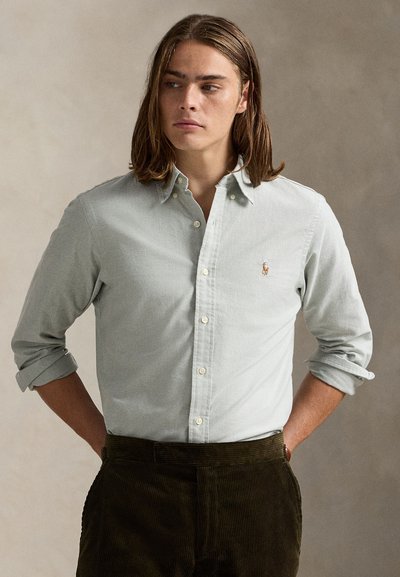 Ανοιχτό πράσινο πουκάμισο με γιακά και μικρό κεντητ ό έμβλημα. Φορεμένο με σκούρο παντελόνι κορδονέ, με ανασηκωμένα μανίκια.