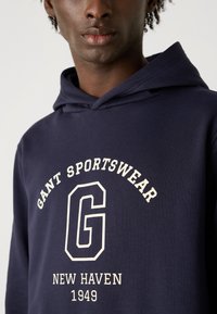 Felpa con cappuccio blu navy in tessuto morbido, presenta una grafica frontale con "GANT SPORTSWEAR," una grande "G," e "NEW HAVEN 1949" in crema chiara.