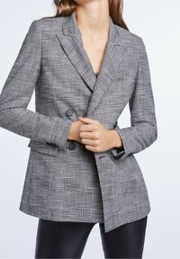 Blazer grigio a scacchi con design doppiopetto, rever a punta e bottoni neri. Indossato sopra pantaloni in pelle nera.