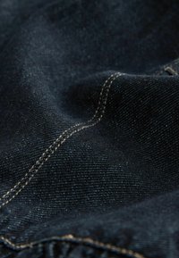 Dunkelblauer Denim-Stoff mit einer strukturierten Oberfläche, die subtile gewebte Muster aufweist und mit gelben Nähten entlang der Nähte sichtbar ist.