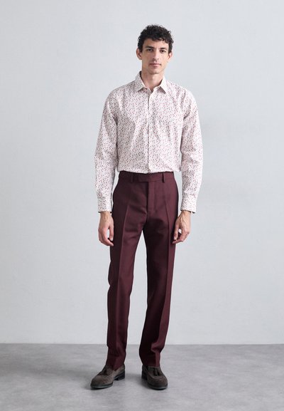 White shirt with small red and blue floral patterns, paired with dark maroon trousers. Model wears brown shoes, stands against a neutral background.