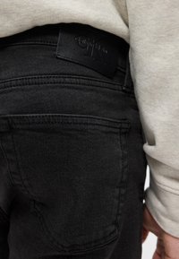 Πίσω τσέπη μαύρου τζιν με μαύρο λογότυπο Calvin Klein, φορεμένα με ανοιχτό μπεζ μακρυμάνικη μπλούζα.