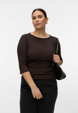 Kvinde med mørkt hår samlet i en hestehale iført sort langærmet skjorte og bukser, der holder en sort skuldertaske mod en hvid baggrund.