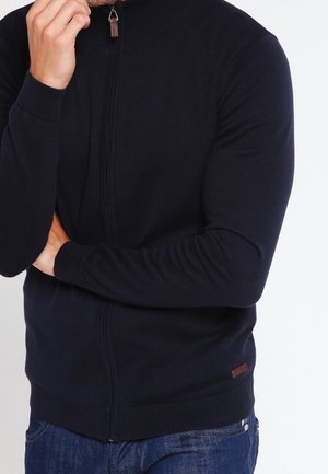 Mann trägt marineblauen Reißverschluss-Pullover und blaue Jeans, eine Hand berührt das Kinn, der andere Arm ist verschränkt.