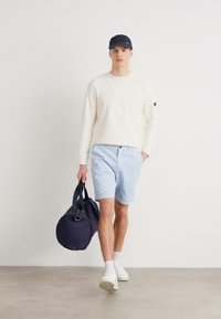 Sweatshirt creme, calças de jogging azul claro e ténis brancos. A carregar uma mala de desporto azul-marinho. Design simples; roupa casual de verão.