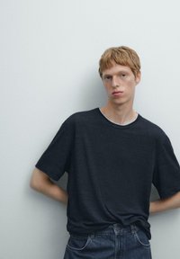 Tmavě šedé tričko s krátkým rukávem a kulatým výstřihem, vyrobené z jemné tkaniny, nošené s modrými džínami. Jednoduchý design, bez viditelných vzorů nebo grafik.