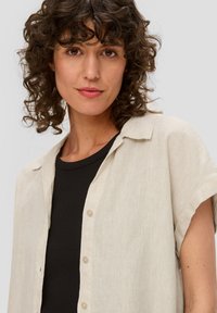 Vrouw met krullend bruin haar, gekleed in een beige overhemd met korte mouwen over een zwarte top, kijkt rechtstreeks naar de camera.