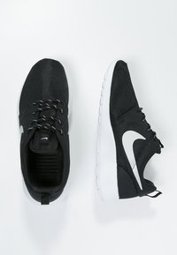 Une paire de baskets noires avec des semelles blanches, un swoosh Nike blanc sur le côté et des lacets noir et blanc sur un fond blanc.