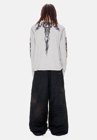 Άτομο με μακριά dreadlocks που φοράει ανοιχτό γκρι μακρυμάνικο πουκάμισο με μαύρα φυλετικά σχέδια και χαλαρό μαύρο cargo παντελόνι, στέκεται με την πλάτη προς τα εμπρός.