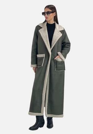 Lungo cappotto verde oliva con colletto e rifiniture in faux shearling crema, tasche laterali e vestibilità ampia; modello che indossa un top nero e occhiali da sole.
