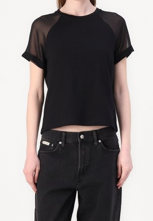 Nainen, jolla on lyhythihainen musta paita, jossa läpikuultavat olkapäät, ja mustat korkeavyötäröiset farkut, joissa näkyy brändilappu.