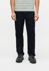 Άνδρας που φοράει μαύρο παντελόνι με ίσια γραμμή, μαύρα και γκρι αθλητικά παπούτσια, και λευκό πουκάμισο με πράσινες ρίγες, στέκεται μπροστά σε λευκό φόντο.