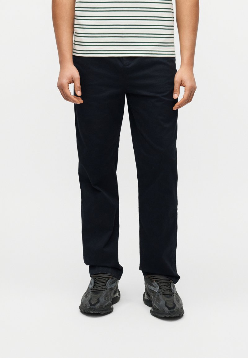 Άνδρας που φοράει μαύρο παντελόνι με ίσια γραμμή, μαύρα και γκρι αθλητικά παπούτσια, και λευκό πουκάμισο με πράσινες ρίγες, στέκεται μπροστά σε λευκό φόντο.