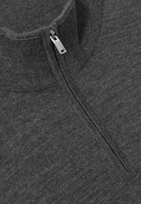 Close-up af en mørkegrå strikket sweater med høj krave og en lille sølvlynlås, der delvist er lynet i toppen.