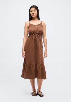Žena stojí, má na sebe hnedé midi šaty bez rukávov s gombíkmi, opaskom v páse, volánikový lem a tmavé rovné topánky na remienky.