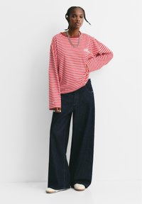 Donna che indossa una camicia a maniche lunghe rossa e bianca a righe, jeans larghi scuri, scarpe bianche e collane di catena argentata, in piedi con una mano sul fianco.