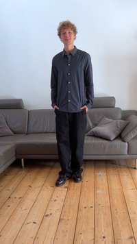 Svart skjorte med knapper, avslappet passform, kombinert med løse svarte bukser. Gulvet er laget av lyse treplanker; sofaen er grå med teksturerte puter.