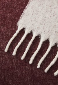 �Écharpe en laine blanche avec des bords à franges, présentée sur un fond bourgogne foncé ; texture douce avec un léger motif de taches.