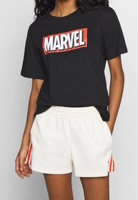 Person iført en sort Marvel-logo t-shirt stoppet i hvide shorts med røde striber på siden, hænderne i lommen, neutral baggrund.