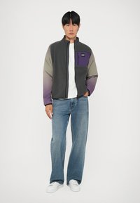 Fleece striukė su aukštu apykakle, pilkai violetinėmis gradientinėmis rankovėmis ir priekinėmis kišenėmis, kuriose yra logotipas. Derinama su šviesiai mėlynomis džinsinėmis kelnėmis ir baltomis sportbačiais.