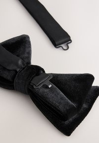 Nœud papillon en velours noir avec une bande en satin lisse et un détail de boucle métallique. Le tissu est doux et le design présente une forme classique de papillon.