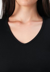 Nahaufnahme des Halses und oberen Brustbereichs einer Person, die ein schwarzes V-Ausschnitt-Oberteil trägt, wobei die glatte Haut und die Textur des Ausschnitts hervorgehoben werden.