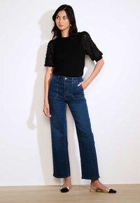 Must have lühikeste varrukatega must topp, millel on pitsdetailid, koos kõrge taljega siniste denim laiekeste pükstega. Mudel kannab kuldseid ja musti madalaid kingi.
