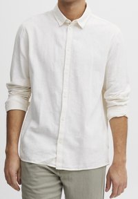 Chemise en lin couleur crème avec un col boutonné, manches longues retroussées et un ourlet droit. Caractérisée par une texture subtile et six boutons.