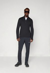 Maglia nera con zip e maniche lunghe abbinata a pantaloni sportivi grigio scuro. Caratteristiche includono vestibilità slim, texture liscia e elementi di design pratici.