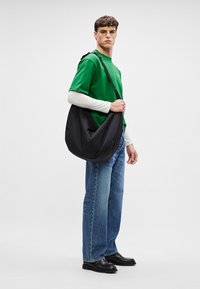 Saco de ombro de tecido preto com forma curva. Modelo veste uma t-shirt verde, uma camisa de manga longa clara, jeans azuis e sapatos estilo loafers pretos.