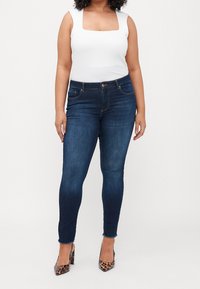 Dark blue skinny jeans with a high waist, featuring slight fading, front pockets, and a frayed hem paired with a white square-neck top.