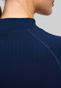 Granatowa sportowa bluzka z wysokim kołnierzem, wykonana z teksturowanego materiału z drobnymi otworkami przypominającymi dziurki i kontrastowymi szwami dla dodatkowego efektu wizualnego.