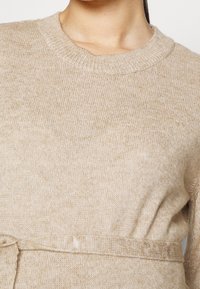 Beige pletený svetr s kulatým výstřihem a texturovaným povrchem. Má odpovídající pásek v pase pro zvýraznění detailu.