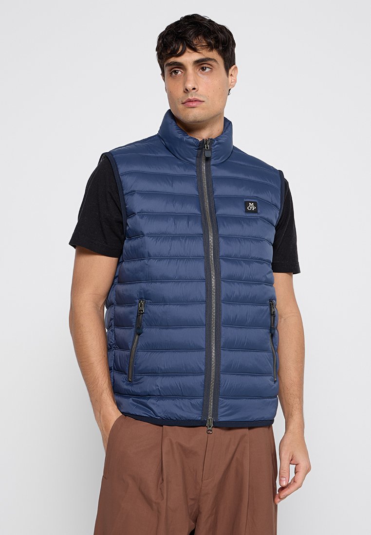 Granatowa, pikowana kamizelka puchowa z wysokim kołnierzem, z zamkiem z przodu oraz dwoma bocznymi kieszeniami na zamek. Nosić na czarnej koszuli.