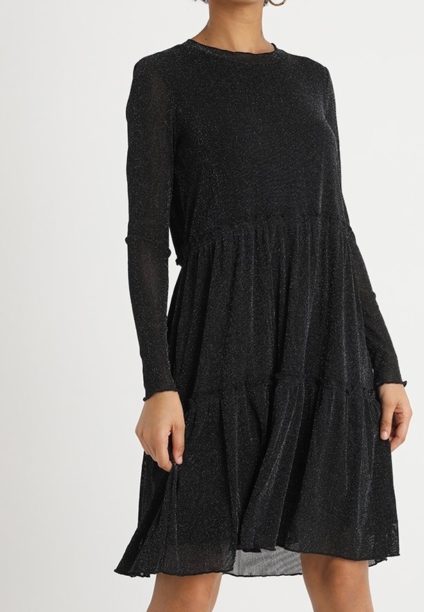 Femme portant une robe noire à manches longues, longue jusqu'aux genoux, avec un léger éclat et des volants froncés sur la jupe.