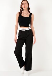 Črn krojeni top z širokimi naramnicami, v kombinaciji z visokimi črnimi hlačami, ki imajo bel pas v črtah. Model nosi bele superge.