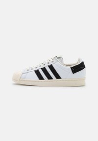 White sneaker with three black stripes, rubber toe cap, and beige sole. Features lace-up design and a textured back patch.
