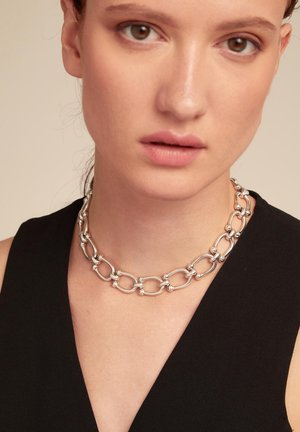 Collar de cadena de plata con eslabones ovalados y acentos redondos, sobre una blusa negra. Acabado suave y pulido con un diseño interconectado.