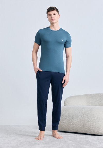 Homme debout pieds nus à l'intérieur portant un t-shirt ajusté turquoise et un pantalon de jogging bleu marine à côté d'un canapé blanc texturé sur un tapis gris.