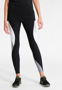 Person med svart, vit och grå färgblockerade leggings och svarta Nike-sneakers står på en vit bakgrund.