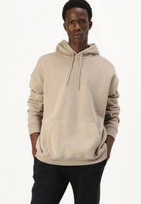 Bežs hoody mīkstā audumā ar priekšējo kabatu, regulējamu kapuci ar saiti un ribotu piedurkņu galiem, savienots ar melnajām biksēm.